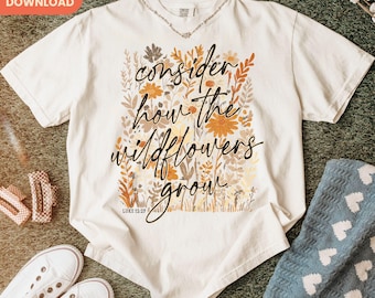 Puede incluir: Camiseta color crema con la frase "consider how the wildflowers grow" en escritura negra, rodeada de ilustraciones florales en tonos marrón, naranja y amarillo. El texto "Luke 12:27" está debajo. Se ve un collar plateado. Ropa casual.