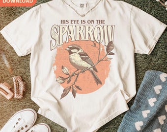 Puede incluir: Camiseta de color crema con un gorrión posado en una rama, sobre un círculo color melocotón. El texto "HIS EYE IS ON THE SPARROW" está arqueado sobre el pájaro. La camiseta se combina con zapatillas blancas y una manta azul.