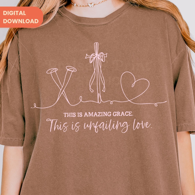 以下が含まれることがあります： 十字架、釘、ハートのピンク色の線画が描かれた茶色のTシャツ。「THIS IS AMAZING GRACE」と「This is unfailing love」の文字が絵の下にあります。シャツにはデジタルダウンロードタグが付いています。