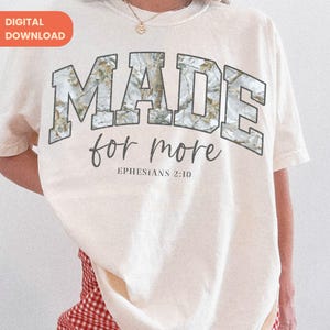 Op de afbeelding: Cr&egrave;mekleurig T-shirt met de woorden "MADE for more" in een camouflage-stijl lettertype. Onder de tekst staat de zin "Ephesians 2:10" in een kleiner lettertype. Het shirt heeft een relaxte pasvorm.