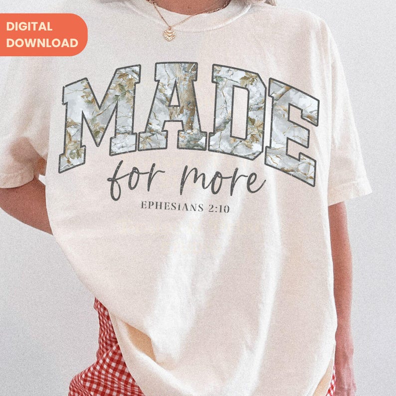 Op de afbeelding: Cr&egrave;mekleurig T-shirt met de woorden "MADE for more" in een camouflage-stijl lettertype. Onder de tekst staat de zin "Ephesians 2:10" in een kleiner lettertype. Het shirt heeft een relaxte pasvorm.