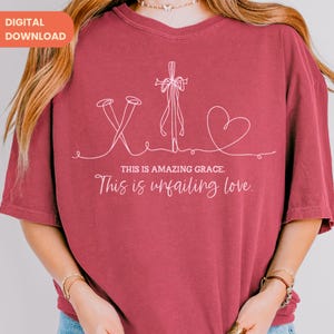 以下が含まれることがあります： 釘、十字架、ハートが描かれた白いグラフィックデザインのマルーンTシャツ。「THIS IS AMAZING GRACE. This is unfailing love.」の文字がプリントされています。ゆったりとしたフィット感のシャツです。