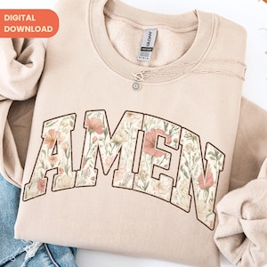 Op de afbeelding: Beige sweatshirt met het woord "AMEN" in een bloemenontwerp. De letters zijn gevuld met een patroon van roze, perzik en crèmekleurige bloemen en groene bladeren. Een delicate zilveren ketting met een hanger is over de sweatshirt gedrapeerd.