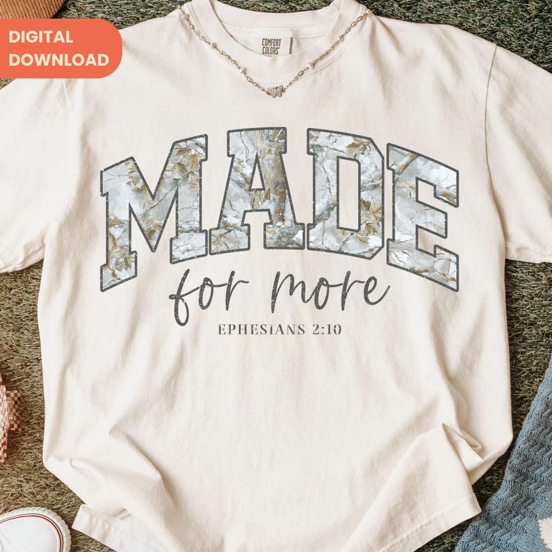 Op de afbeelding: Cr&egrave;mekleurig T-shirt met de woorden "MADE for more" in een grijze lettertype in collegestijl. Het woord "MADE" heeft een camouflagepatroon. De tekst "Efezi&euml;rs 2:10" staat onder de zin. Een digitaal downloadlabel staat in de linkerbovenhoek.
