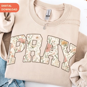 May include: A beige sweatshirt with the word "PRAY" in large, floral-filled letters. The floral design includes pink, yellow, and green elements. A delicate silver necklace with a pendant is draped across the sweatshirt.