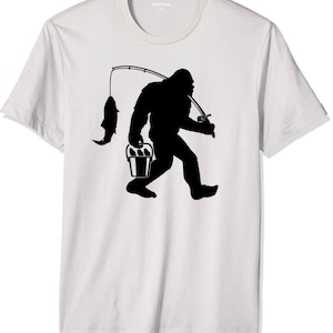 May include: A white t-shirt with a black silhouette of Bigfoot holding a bucket of fish and a fishing rod.