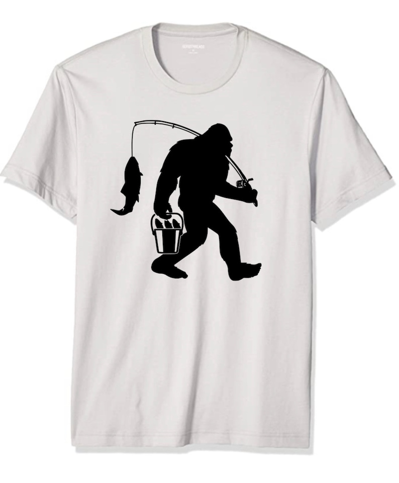 May include: A white t-shirt with a black silhouette of Bigfoot holding a bucket of fish and a fishing rod.