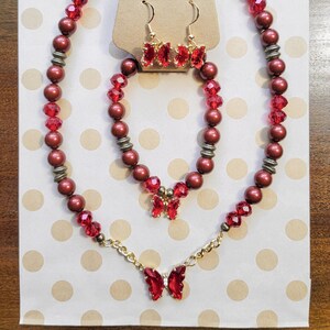 Könnte beinhalten: Ein Set aus rotem und goldenem Schmuck mit Schmetterlingsanhängern. Das Set enthält eine Halskette, ein Armband und Ohrringe. Der Schmuck ist handgefertigt mit Liebe.