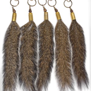 May include: Five keychains with brown and gray fur tails. Each keychain has a brass-colored clasp, ring, and a decorative bead. The fur tails are attached to brass-colored bullet casings.