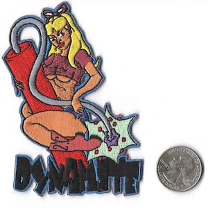 May include: An embroidered patch featuring a cartoon woman in a red and white outfit sitting on a dynamite stick with the word "Dynamite" written below. The patch is approximately the size of a US quarter.