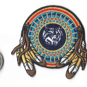 May include: Embroidered patch featuring a circular design with a wolf in the center, surrounded by a blue and gold pattern. The patch includes brown and yellow feathers on each side. A quarter coin is next to the patch for size reference.
