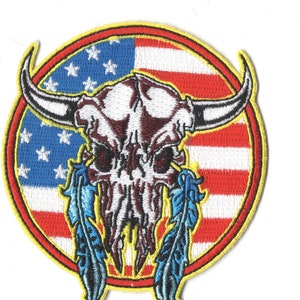 May include: A round embroidered patch with a white bull skull with red and black details, two blue feathers, and an American flag design in the background. The patch is surrounded by a yellow border.