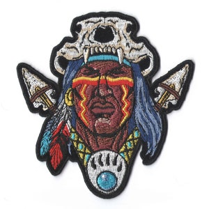 May include: Embroidered patch featuring a Native American chief with a bear skull headdress. The patch has a detailed design with vibrant colors, including red, blue, and yellow. Two arrowheads flank the chief's face, and a bear claw with a blue gem is at the bottom.