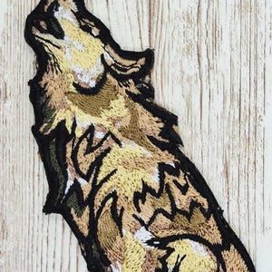 May include: Embroidered patch featuring a howling wolf. The patch showcases a detailed design with shades of tan, brown, and black, creating a textured appearance. The wolf is depicted in a dynamic pose, with its head raised and mouth open.
