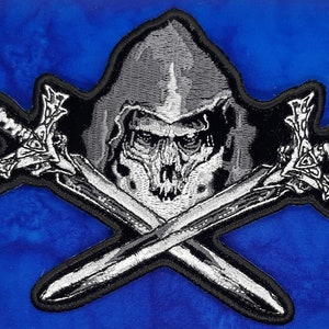 May include: Embroidered patch featuring a skull wearing a hood, with two crossed swords. The design is primarily black and white, with detailed stitching. The patch has a black border and is set against a blue background.