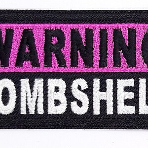 May include: Black embroidered patch with white text that reads "WARNING BOMBSHELL" and a pink background for the word "WARNING".