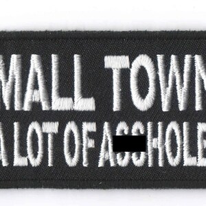 May include: Black rectangular patch with white embroidered text: "FOR A SMALL TOWN IT SURE HAS A LOT OF..." A quarter coin is next to the patch. The patch is likely for a jacket or bag.