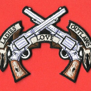May include: Embroidered patch with two crossed revolvers, a banner reading "LADIES LOVE OUTLAWS" in white lettering, and brown and white accents.