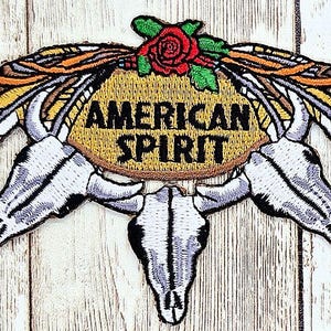 May include: Embroidered patch featuring the words "AMERICAN SPIRIT" in black on a tan oval. The design includes three white and gray bull skulls, feathers, and a red rose, with orange and yellow accents.