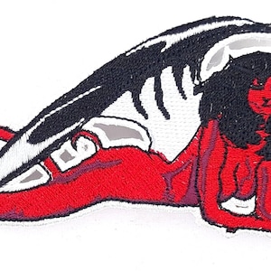 May include: Embroidered iron-on patch featuring a red, winged, female figure with black hair and horns. The figure is lying on its side with its wings spread out behind it.