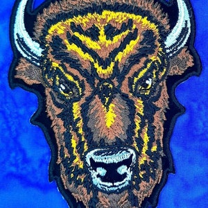 May include: Embroidered patch featuring a detailed bison head. The patch showcases a brown, yellow, and black color palette with white horns and a blue background. The design is intricately stitched, creating a textured appearance.