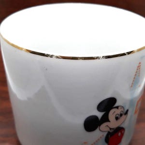 K&ouml;nnte beinhalten: Wei&szlig;e Keramiktasse mit goldfarbenem Rand und einem Comicbild von Mickey Mouse. Die Tasse hat ein blaues und oranges Design.