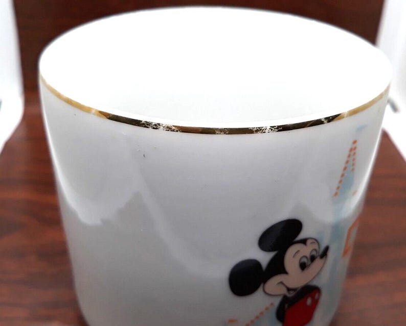 K&ouml;nnte beinhalten: Wei&szlig;e Keramiktasse mit goldfarbenem Rand und einem Comicbild von Mickey Mouse. Die Tasse hat ein blaues und oranges Design.