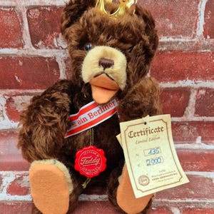 May include: A brown teddy bear with a gold crown, wearing a red and white sash that says "ein Berliner". The bear has a tan snout and paws, and a red "Hermann Teddy Original" seal. A certificate is attached.