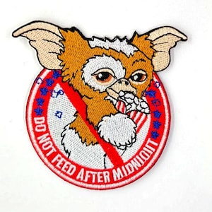 May include: A circular embroidered patch featuring a brown and white creature with large ears holding a striped object. A red diagonal line crosses the image over a red border with blue stars and the text DO NOT FEED AFTER MIDNIGHT.