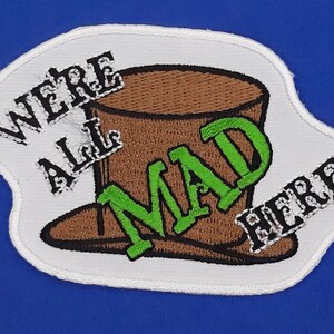 May include: White embroidered patch with a brown top hat and the text "We're All Mad Here" in black and green lettering.