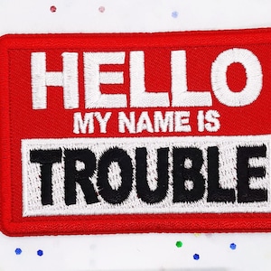 May include: Red and white embroidered patch with the text "HELLO MY NAME IS TROUBLE".