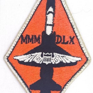 May include: A diamond-shaped patch with a white outline. The patch is orange with a black silhouette of a jet plane with white wings. The text "MMM DLX" is embroidered in black on the patch.