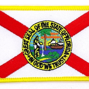 May include: A white embroidered patch with a red diagonal cross and the state seal of Florida. The seal is a yellow circle with the words "Great Seal of the State of Florida" and "In God We Trust" around the edge. The seal depicts a sun, a tree, a river, and a Native American man.