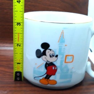 K&ouml;nnte beinhalten: Wei&szlig;e Keramiktasse mit goldfarbenem Rand, die Mickey Mouse vor einer blauen und wei&szlig;en Illustration von Schloss Aschenputtel zeigt. Mickey Mouse tr&auml;gt rote Shorts und gelbe Schuhe.