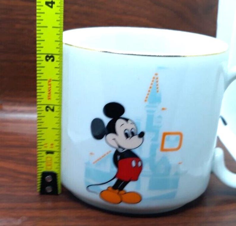 K&ouml;nnte beinhalten: Wei&szlig;e Keramiktasse mit goldfarbenem Rand, die Mickey Mouse vor einer blauen und wei&szlig;en Illustration von Schloss Aschenputtel zeigt. Mickey Mouse tr&auml;gt rote Shorts und gelbe Schuhe.