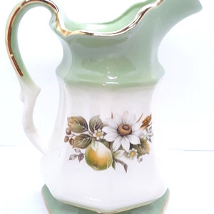 May include: A white ceramic creamer with a green top and gold trim. The creamer features a floral design with a pear and a daisy.