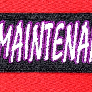 May include: Black fabric patch with white lettering that reads "HIGH MAINTENANCE" in a stylized font. The lettering is outlined in purple.