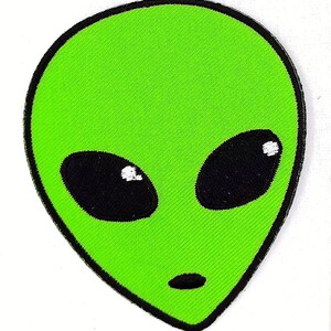 May include: Green alien face with black eyes and a white mouth. The alien face is a cartoon style and is embroidered on a fabric patch.