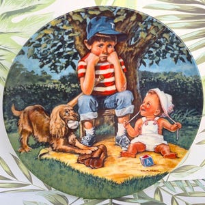May include: A vintage ceramic plate featuring a painting of a boy sitting on a bench with a dog and a baby girl. The boy is wearing a blue baseball cap and a red and white striped shirt. The dog is brown and white and is holding a baseball in its mouth. The baby girl is wearing a white dress and a pink hat. The plate has a green border and a white background.