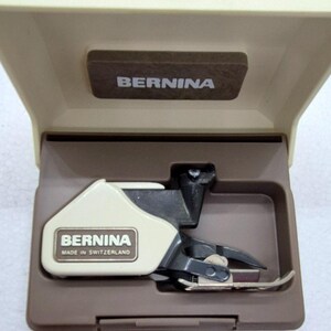 May include: A Bernina sewing machine accessory, featuring a cream-colored foot with black and silver components, housed in a brown case. The case lid displays the brand name "BERNINA". The accessory is labeled "BERNINA MADE IN SWITZERLAND".