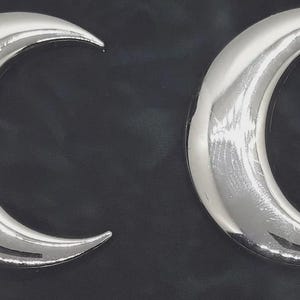 May include: Two silver crescent moon-shaped objects. The moons have a smooth, reflective surface and are set against a dark, textured background. The design is simple and elegant, with a modern aesthetic.