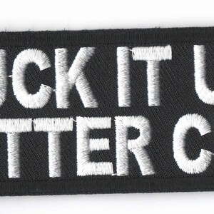 May include: Black rectangular embroidered patch with white text that reads "SUCK IT UP BUTTER CUP". The patch has a black border and is likely designed to be sewn or ironed onto clothing or accessories.