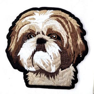 May include: Embroidered patch featuring a Shih Tzu dog's face. The patch has a black border and showcases the dog's brown, white, and tan fur. The dog's eyes and nose are detailed with dark brown thread, creating a realistic appearance.
