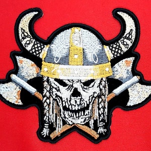 May include: Embroidered patch featuring a skull wearing a horned Viking helmet with crossed axes behind it. The skull is white with black eyes and a black mouth. The helmet is gray with gold trim. The axes are silver with black handles.