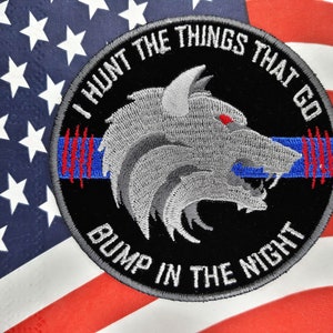 May include: A black and gray embroidered patch with a wolf's head and the text "I HUNT THE THINGS THAT GO BUMP IN THE NIGHT". The patch has a blue and red stripe across the middle.
