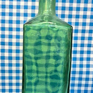 May include: A tall, green glass bottle with a rectangular body and a long, narrow neck. The bottle has a slightly textured surface and is set against a blue and white checkered background. The bottle's height is approximately 10 inches.
