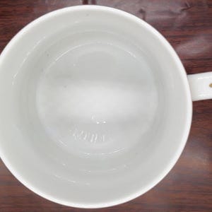 K&ouml;nnte beinhalten: Wei&szlig;e Keramiktasse mit goldenem Henkel. Die Tasse ist innen leer.