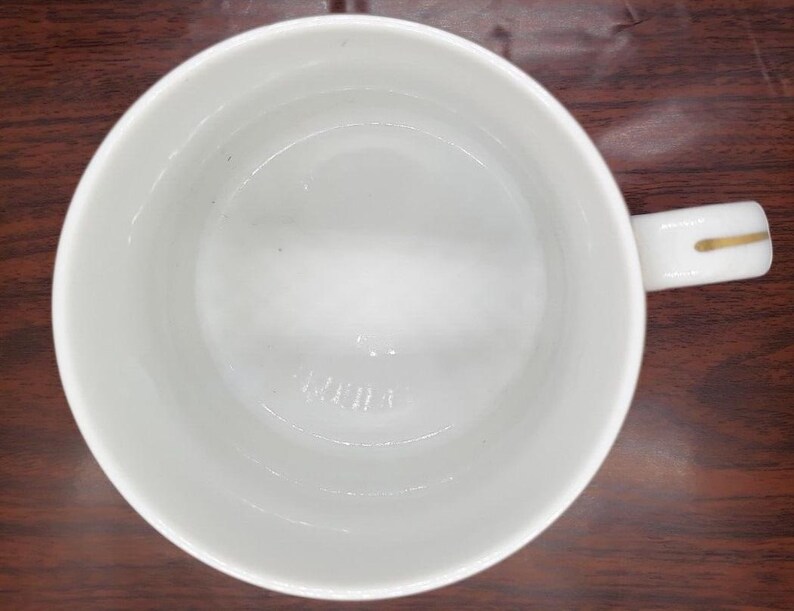 K&ouml;nnte beinhalten: Wei&szlig;e Keramiktasse mit goldenem Henkel. Die Tasse ist innen leer.