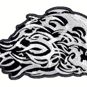 May include: Embroidered patch featuring a stylized skull design with intricate white detailing against a black background. The patch has a flame-like shape, with a black border. Ideal for jackets, bags, or other apparel.