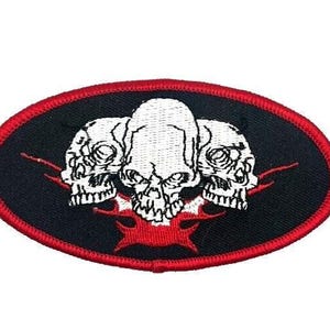 May include: An oval patch featuring three white skulls on a black background, framed by a red border. Red flame-like shapes emanate from the bottom of the skulls. The patch is likely for clothing or accessories.